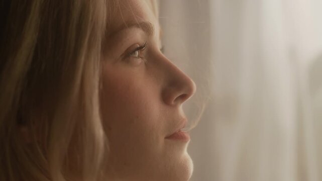 Side close up shot of young adult Caucasian woman with blonde hair contemplating standing by window with breeze blowing in morning sunlight
