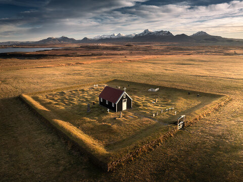 Aerial view of the historic Budakirkja black church surrounded by a stone wall in a vast grassy field with snow-capped mountains in the background Iceland.
