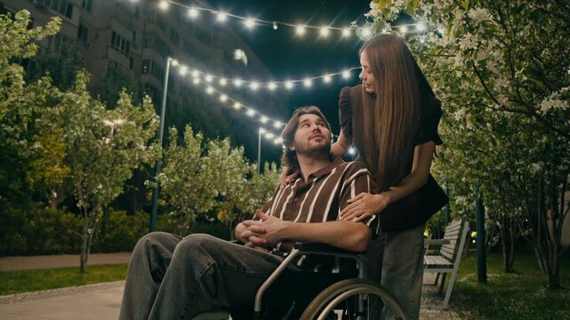 Man in wheelchair and woman connect during relaxed date in illuminated park. Natural greenery and blossoms create peaceful accessible scene.