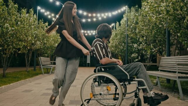 Woman pushes wheelchair user in joyful circle while laughing in park. Blooming trees and string lights frame cheerful lifestyle moment.