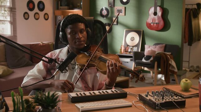 Medium portrait shot of young adult Black male musician with headphones on pausing from playing violin, smiling at camera while recording music in equipped home studio