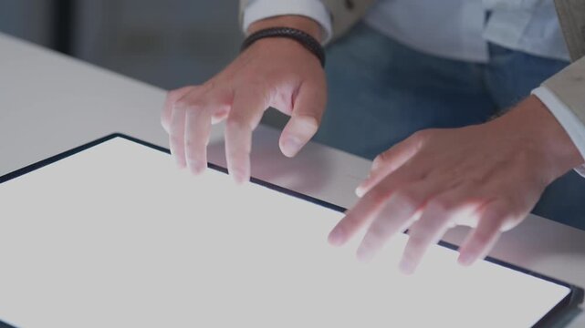 Close up view of a tech specialist hands typing on a blank white digital tablet screen layout on the desk during a late night corporate planning session.