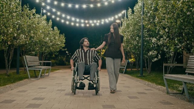 Young couple walks together through park under glowing string lights at night. Woman guides wheelchair user along peaceful romantic path.
