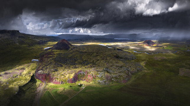 Aerial view of red volcanic cones and rugged lava fields under dramatic storm clouds with shafts of sunlight Iceland.