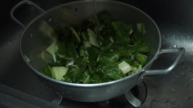 High angle view of hands washing chopped fresh chard leaves in a metal colander inside a sink
