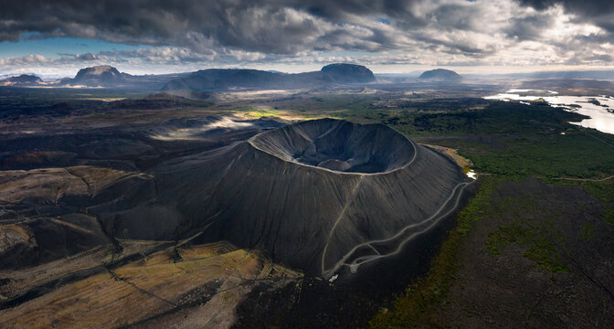 Aerial view of the massive Hverfjall volcanic crater with its dark ash slopes and circular rim under a dramatic cloudy sky in the volcanic landscape of Iceland.