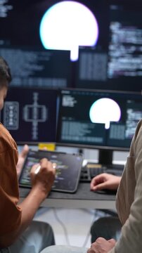 Over the shoulder view of tech developers analyzing software coding lines and circuit architectures across dual monitor desktop screens in a startup office.