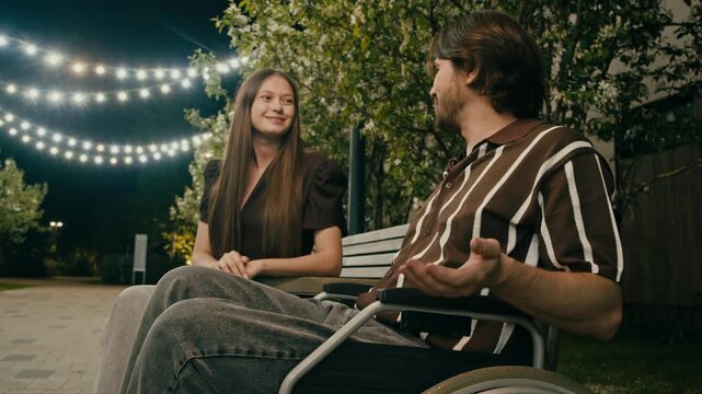Young woman talks with wheelchair user in park under glowing string lights. Warm evening setting creates tender romantic conversation.