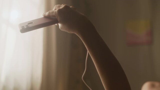 Tilt down handheld shot of dreamy Caucasian girl taking selfies on smartphone while relaxing in bed under morning sunlight