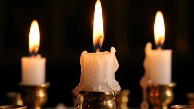 Close-up of three burning candles with flickering flames. Melted wax drips down vintage brass candlesticks against a black background. Spiritual and remembrance concept