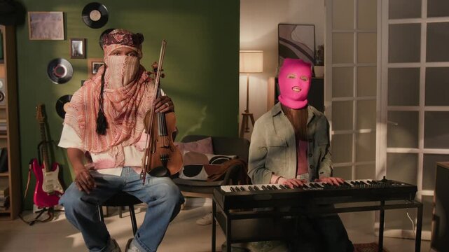 Portrait of young adult Black male violinist wearing shemagh posing in home studio next to female synth player covering face with pink balaclava, slow motion