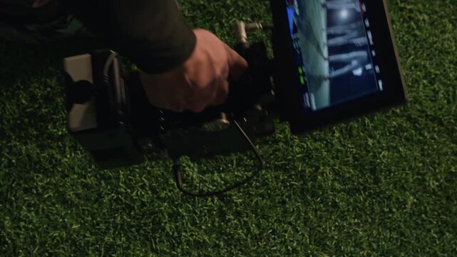 Close-up lockdown shot of hand of anonymous male operator lifting up camera from green turf while shooting sports show at stadium, legs of football or rugby team players visible on screen