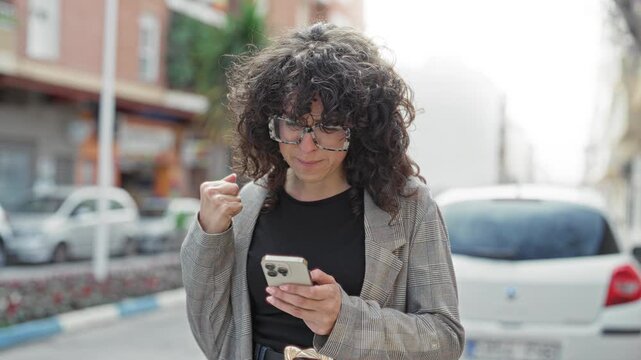 Young woman with curly hair and patterned jacket holding a smartphone, making a clenched fist pump while checking the screen on a city street; joy.