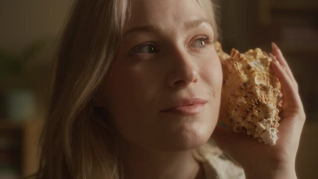 Close up shot of dreamy blonde Caucasian woman holding big shell to ear and imagining ocean sound while relaxing at home under natural light