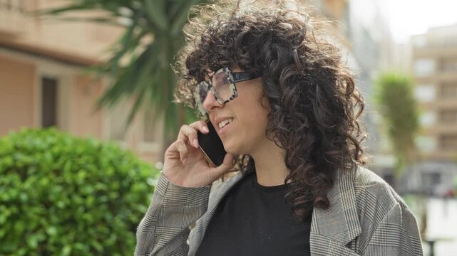 Young woman with curly hair and tortoiseshell glasses holds smartphone to her ear smiling while talking on a city street near palm plants and apartment buildings; casual contentment.