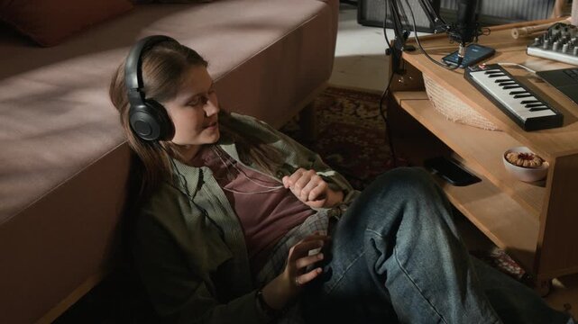 High angle shot of young adult female musician wearing headphones enjoying listening to recorded takes in home studio while checking quality of instrumental performance