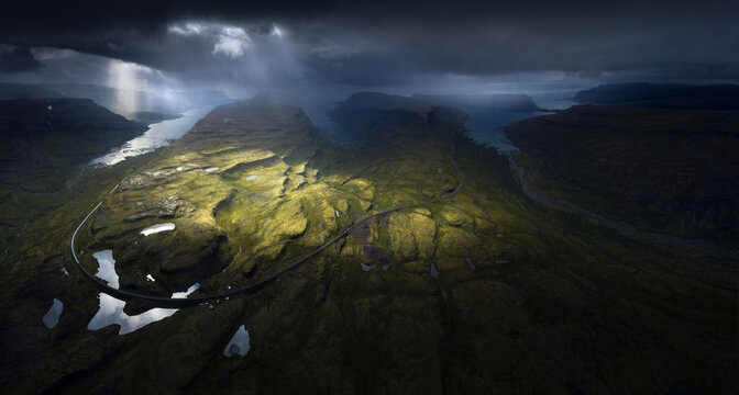 Aerial view of a winding road through green mossy hills under dramatic sunbeams and dark storm clouds in Iceland.