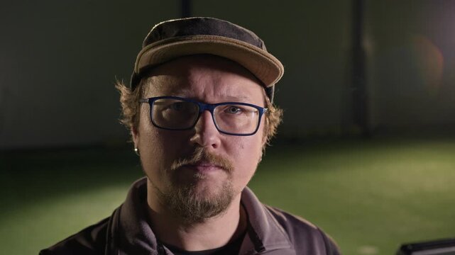 Close-up face portrait of confident mature Caucasian male TV show director filming sporting event at arena or stadium, turning and looking at camera, wearing glasses and baseball cap