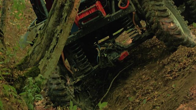 Close up of red off road vehicle climbing steep, muddy hill in woods. Low angle shot highlights powerful suspension, axle, and large tires gripping difficult terrain. Action packed and adventurous