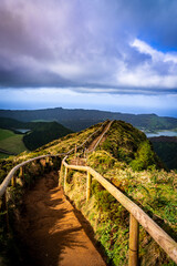Fototapeta premium Wooden-railed hiking trail winding along a lush volcanic ridge in the Azores with crater lakes and ocean views under a dramatic cloudy sky.