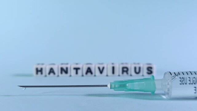 Close-up rack focus from Hantavirus letter blocks to a syringe hypodermic needle tip with a fluid droplet. Visualizes laboratory science breakthroughs and vaccine manufacturing.