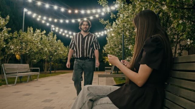 Casual park date begins as man greets woman on wooden bench. Evening lights and natural greenery build relaxed romantic mood.
