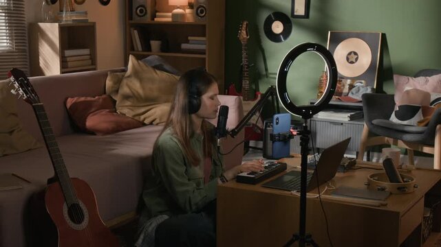 Young adult female multi instrumentalist with headphones performing song while playing MIDI keyboard at music production setup during video recording for social media in home studio