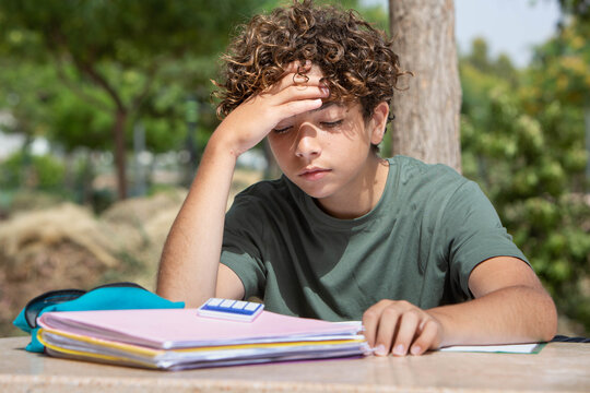 A thoughtful teenage boy studying outdoors in a park, sitting at a table with notebooks and school supplies. The child rests his head on his hand, looking tired or deep in concentration 
