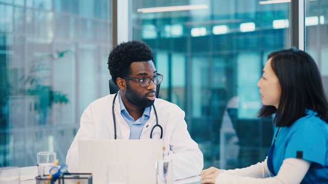 African American male doctor speaking to Asian female colleague during meeting, seated at table with laptop and documents. Medical professionals exchanging insights in bright office.