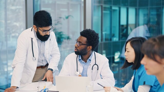 Indian and African American male doctors having discussion around laptop, Asian woman listening, others working. Medical team sharing opinions during collaborative meeting. Multiethnic medics.