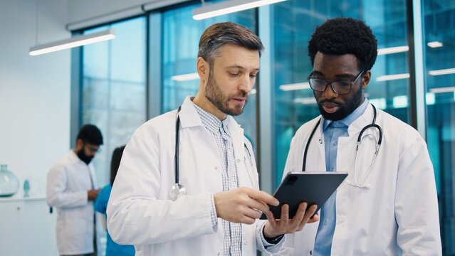 Caucasian male doctor pointing at digital tablet held by African American colleague during discussion in office, both wearing lab coat and stethoscope, collaborating closely. Hospital workday concept.