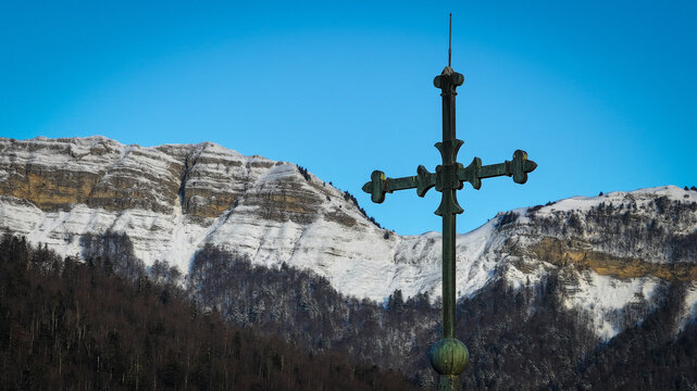 Aerial view of the metal cross of Eglise Saint-Pierre with snow-capped mountains and a clear blue sky in the background Gex, Auvergne-Rhone-Alpes, France.