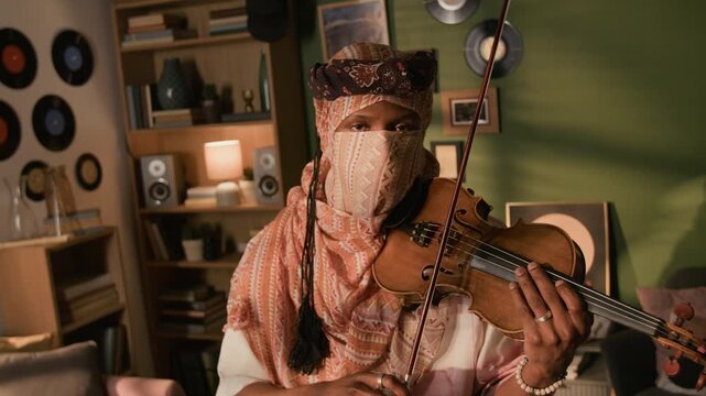 Dutch angle portrait shot of young adult Black male violinist wearing traditional desert clothing performing music on string instrument then facing camera in living room, camera roll movement