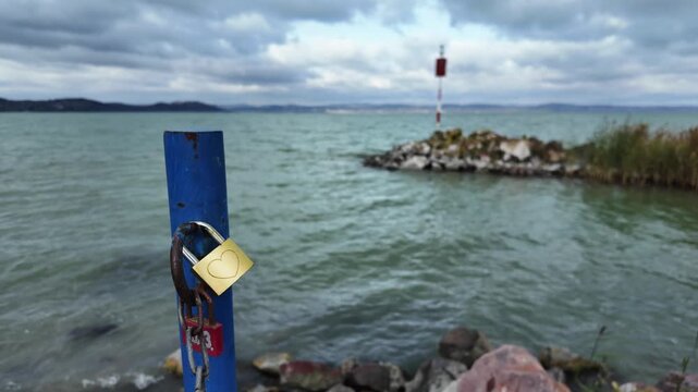 Shiny brass love padlock with engraved heart attached to a post on the shore of Lake Balaton in Zamardi, Hungary. Shallow depth of field love symbol with blurred rocky breakwater in background.
