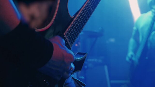 Musician's hands playing electric guitar solo during rock concert. Close up detail of fretboard and strings. Hazy yellow stage lights create energetic, underground vibe. Band performs in background