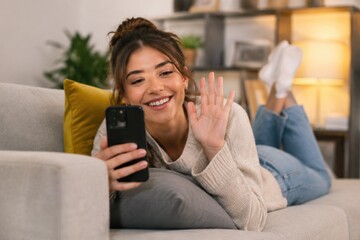 Obraz premium Young woman happily video calling on her smartphone while relaxing on the couch at home