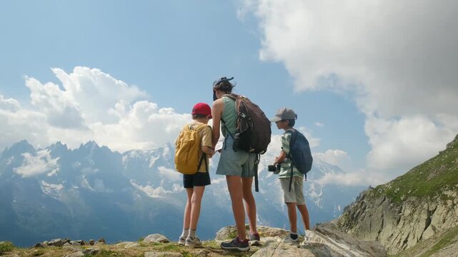 Young mother and her two children standing on an alpine summit, enjoying a panoramic mountain view during a summer hiking adventure and family trekking trip