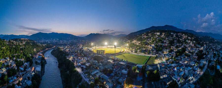 Aerial view of Muzaffarabad Cricket Stadium illuminated at dusk with the surrounding city lights, river, and mountain peaks in Muzaffarabad, Azad Kashmir, Pakistan.