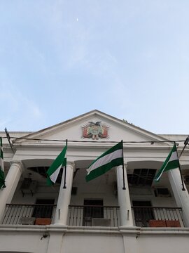Low angle view of the historic government municipal building in Santa Cruz de la Sierra with green and white regional flags and the Bolivian national coat of arms under a clear blue sky.