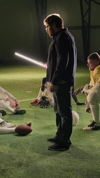 Vertical backstage shot of Caucasian male production crew gaffer holding stick light on set of American football TV show, while team players in jerseys sitting on turf and relaxing after practice