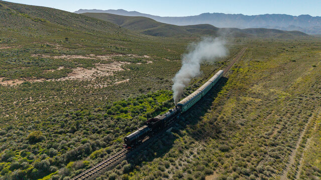 Aerial view of a vintage steam train billowing white smoke as it travels through the rugged semi-arid landscape and rolling hills in Robertson, Western Cape, South Africa.