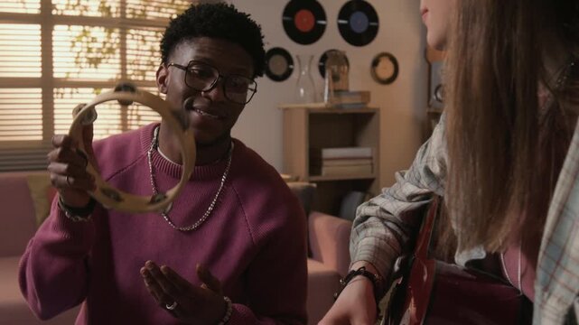 Rack focus shot of young adult female guitarist performing music in duo with African American male musician playing tambourine while improvising together in living room, tilt up camera movement
