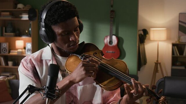 Dolly in shot of young adult Black male musician with headphones on plucking strings of violin while exploring unconventional ways of performance in home based recording studio