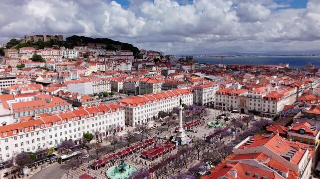 Wide aerial establishing shot over Rossio Square and downtown Lisbon with Castelo de Sao Jorge, jacaranda trees, historic buildings, and the Tagus River visible under dramatic clouds in Portugal.