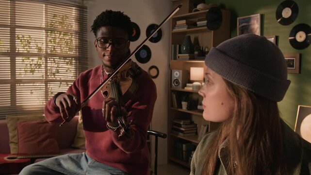Tilt down shot of young adult female musician wearing beanie hat playing synth with Black male violinist accompanying while jamming casually in living room