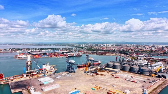 Large commercial port for global trade and logistics. Slow distancing from a maritime facility with cranes, tanks and vessels waiting for the loading and unloading.