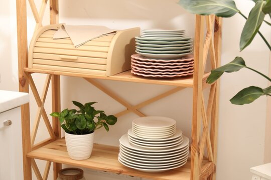 Set of clean plates, plant, breadbox and napkin on wooden shelves in kitchen