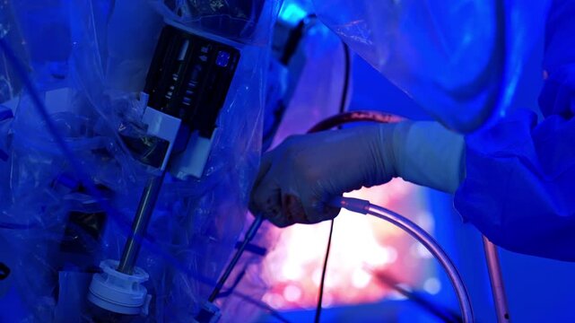 Medic's hand in latex glove hold the surgical device. Robotic arms are coated in plastic nearby. Cooperation of robotic system and human at surgery.