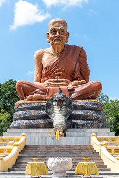 Massive stone statue of a meditating Buddhist monk sitting on a multi-headed cobra, with a smaller monk statue in front, under a clear blue sky.