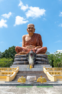 Massive seated monk statue with open-mouthed cobra below, set against a vibrant blue sky with scattered clouds, bathed in clear daylight